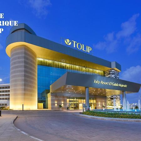 Tolip Resort El Galala Majestic Ain Sukhna Exterior photo The photo shows a modern hotel building named "Tolip Resort & Spa," featuring a sleek architectural design with large glass windows. The hotel is illuminated against a twilight sky, showcasing its prominent signage. In the foreground, there is a well