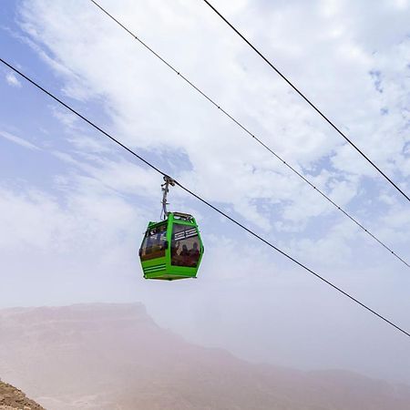 Tolip Resort El Galala Majestic Ain Sukhna Exterior photo The photo shows a green cable car suspended in the air, traveling along overhead cables. The backdrop features a hazy landscape, likely mountainous or elevated terrain, with a cloudy sky. The scene suggests a scenic transportation method, possibly us