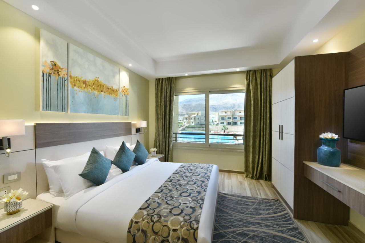 Tolip Resort El Galala Majestic Ain Sukhna Exterior photo The photo shows a modern hotel room. It features a large bed with white bedding and several decorative pillows in shades of teal. Above the bed, there are two pieces of wall art with a landscape theme. The room has a large window with green curtains,