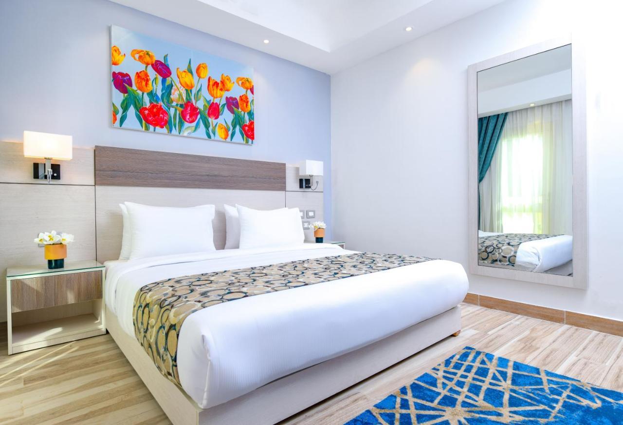 Tolip Resort El Galala Majestic Ain Sukhna Exterior photo The photo shows a modern hotel bedroom. It features a large bed with white linens and a decorative throw blanket. The room has light wooden flooring and is bright, with ample natural light coming from a window. There is a colorful painting of flowers