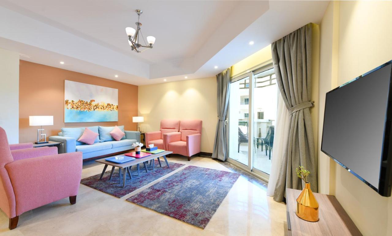 Tolip Resort El Galala Majestic Ain Sukhna Room photo The photo shows a modern living room that features a cozy and stylish design. There is a light blue sofa adorned with pastel pink cushions, accompanied by two pink armchairs. A coffee table sits in front, placed on a colorful area rug that adds warmt