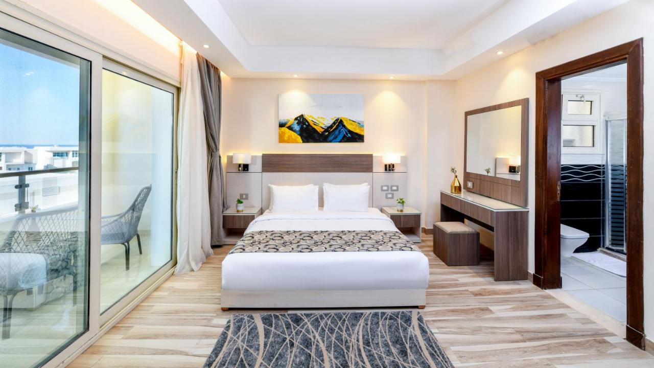 Tolip Resort El Galala Majestic Ain Sukhna Exterior photo The photo shows a modern hotel room featuring a large bed with white linens and a decorative throw. The room has a neutral color palette, accented with warm lighting. There is a piece of artwork above the bed depicting mountains. To the left, there a