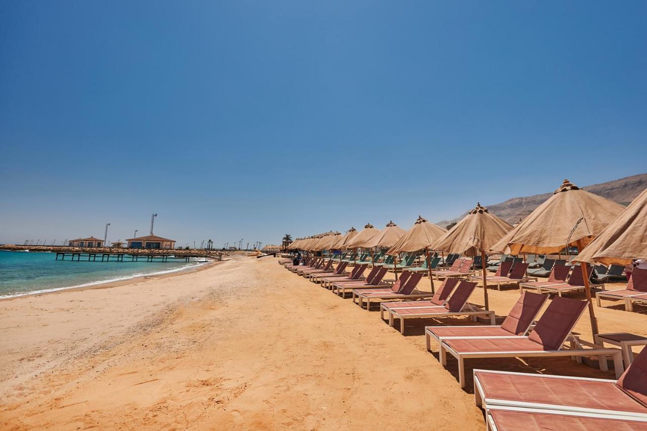 Tolip Resort El Galala Majestic Ain Sukhna Exterior photo The photo shows a scenic beach with a stretch of golden sand under a clear blue sky. There are several beach loungers arranged in rows, each accompanied by thatched umbrellas providing shade. In the background, there is a wooden pier extending into t