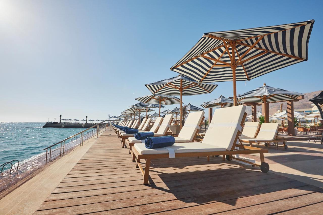 Tolip Resort El Galala Majestic Ain Sukhna Exterior photo The photo depicts a serene beachside setting featuring a row of sun loungers arranged on a wooden deck. Each lounger is equipped with a folded towel and shaded by striped umbrellas. In the background, the water sparkles under bright sunlight, and the