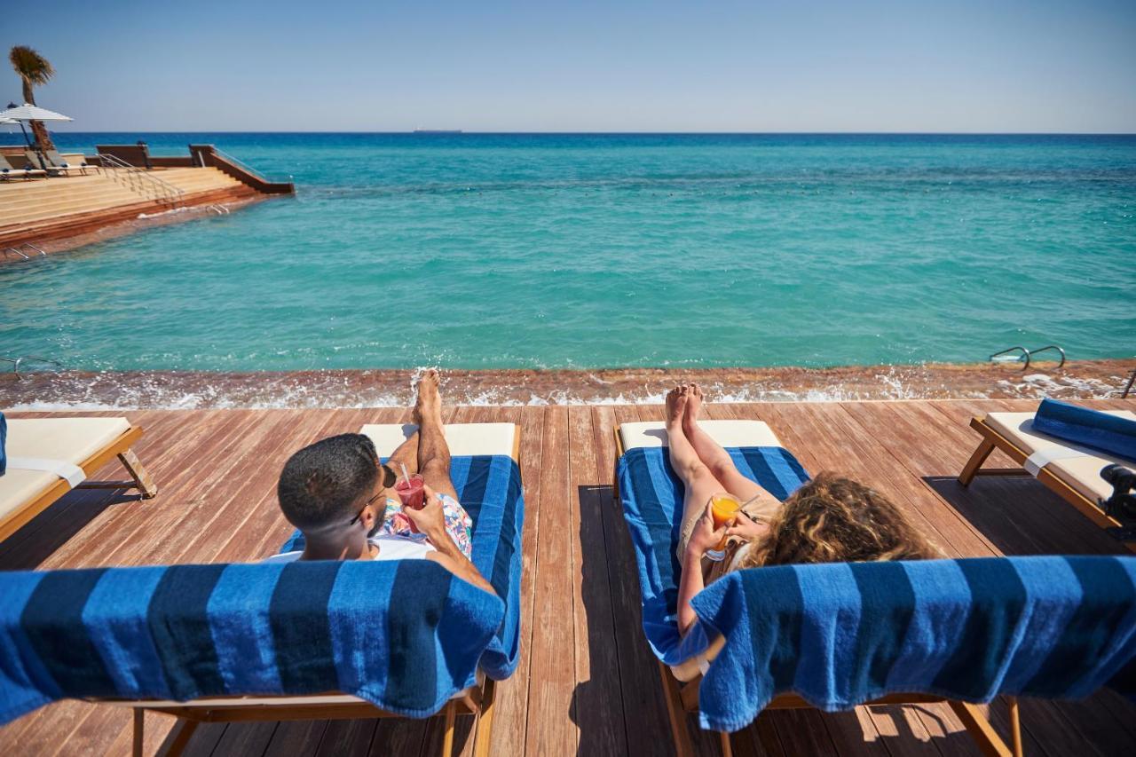 Tolip Resort El Galala Majestic Ain Sukhna Exterior photo The photo depicts two people relaxing on lounge chairs by the waterfront. They are positioned on a wooden deck with a view of the clear, turquoise water. Both individuals are reclining, enjoying the sun, and each has a drink in hand. The scene convey
