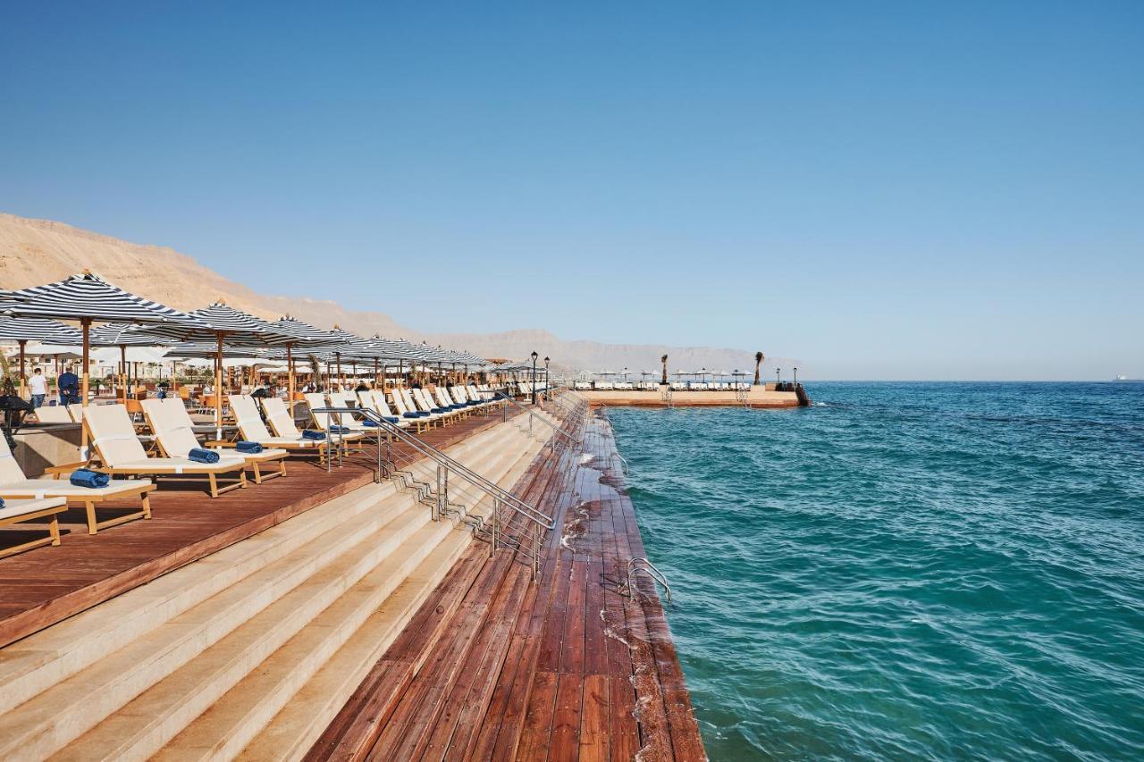 Tolip Resort El Galala Majestic Ain Sukhna Exterior photo The photo shows a seaside area featuring a wooden deck that extends into the water. On the left side, there are several lounge chairs arranged neatly under shade structures, suggesting a relaxing atmosphere. The ocean is visible in the foreground, wi