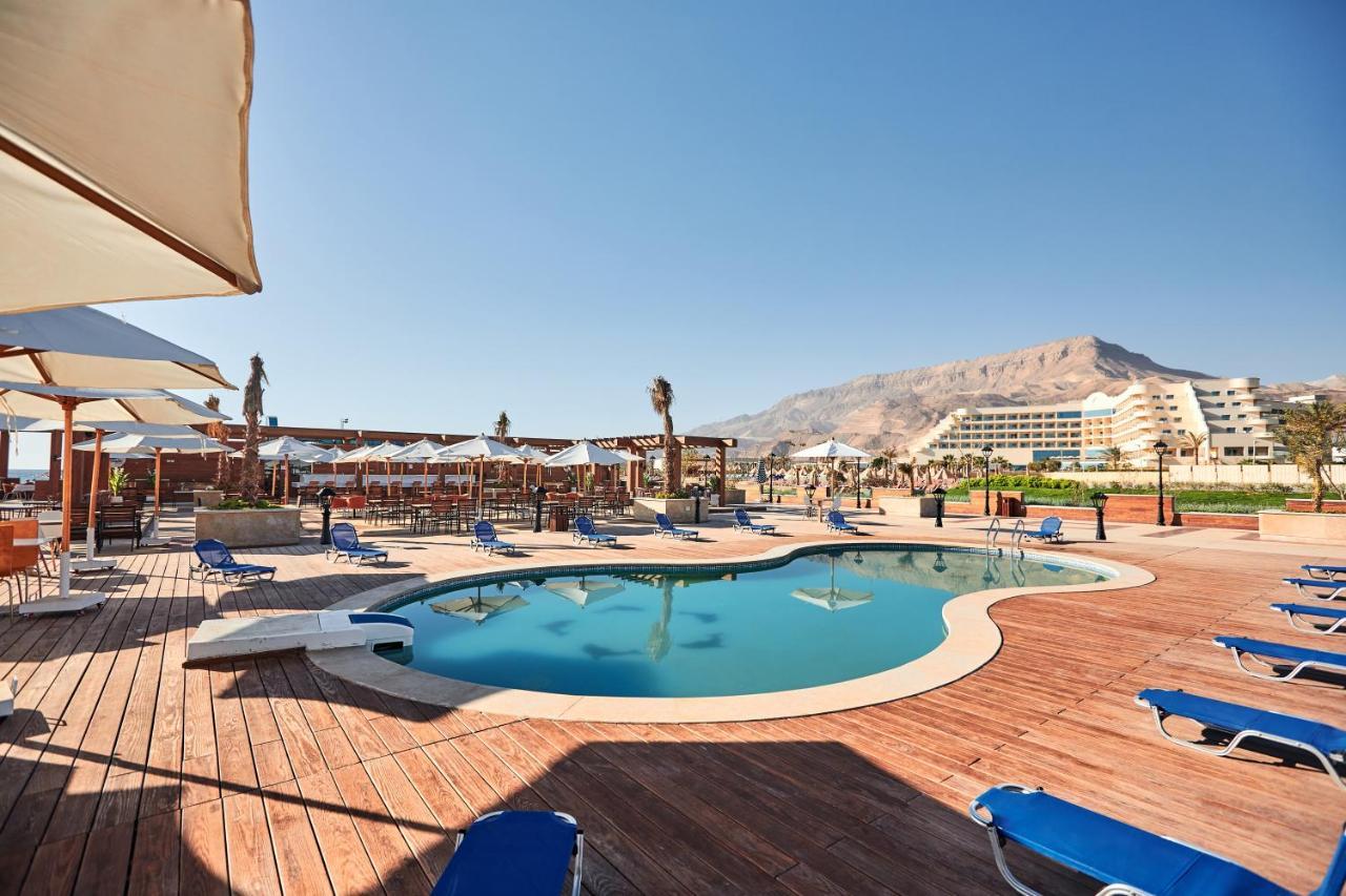 Tolip Resort El Galala Majestic Ain Sukhna Exterior photo The photo shows a swimming pool area surrounded by sun loungers and umbrellas. The pool has a unique, curvy shape and is situated on a wooden deck. In the background, there are several buildings, likely part of a resort or hotel, and a mountainous la