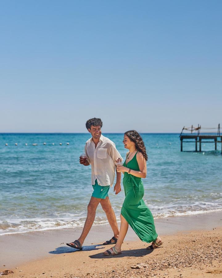Tolip Resort El Galala Majestic Ain Sukhna Exterior photo The photo shows a couple walking along a beach. The man is wearing a white short-sleeved shirt and green shorts, while the woman is dressed in a flowing green dress. They both appear to be enjoying the pleasant weather, with the ocean in the backgrou