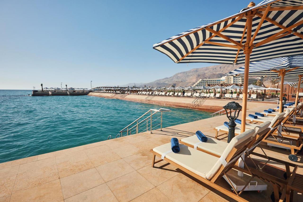 Tolip Resort El Galala Majestic Ain Sukhna Exterior photo The photo shows a scenic beachside area with clear blue water. In the foreground, there are several sun loungers set up on a tiled surface, with blue towels neatly rolled on each one. The loungers are shaded by striped umbrellas. In the distance, a w