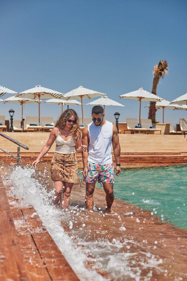 Tolip Resort El Galala Majestic Ain Sukhna Exterior photo The photo depicts a couple walking along a poolside area. They are enjoying a sunny day near a swimming pool, with splashes of water around them. The woman is wearing a light-colored top and a patterned skirt, while the man is dressed in a sleeveless