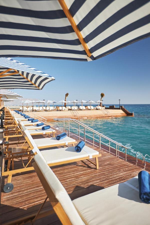 Tolip Resort El Galala Majestic Ain Sukhna Exterior photo The photo depicts a serene outdoor area by the waterfront. There are several lounge chairs arranged in a row, each equipped with blue towels. Large, striped umbrellas provide shade over some of the chairs. In the background, there is a wooden deck th