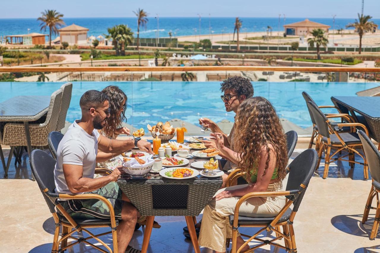 Tolip Resort El Galala Majestic Ain Sukhna Exterior photo The photo shows a group of five people enjoying a meal together outdoors, likely at a restaurant or cafe. They are seated at a table with a beautiful view of a swimming pool and the sea in the background. The table is filled with various dishes, and 