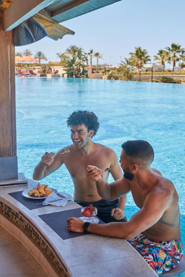 Tolip Resort El Galala Majestic Ain Sukhna Exterior photo The photo features two young men enjoying a meal together. They are sitting at a poolside table with a clear blue swimming pool in the background. The men are shirtless, suggesting a casual, relaxed atmosphere, likely in a warm environment. One man i
