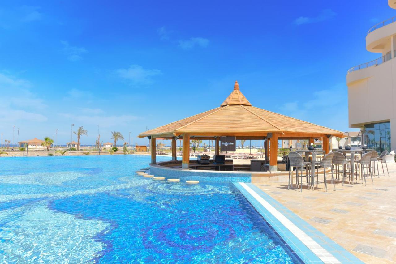 Tolip Resort El Galala Majestic Ain Sukhna Exterior photo The photo shows a beautiful outdoor setting featuring a large swimming pool with clear blue water. In the center, there is a thatched-roof gazebo, which serves as a shaded seating area. Surrounding the gazebo are lounge chairs and a bar area with hig