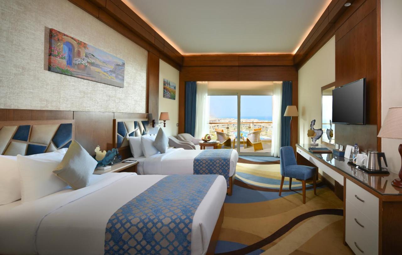 Tolip Resort El Galala Majestic Ain Sukhna Exterior photo The photo shows a well-decorated hotel room featuring two beds with decorative pillows. The room has wooden accents and modern furnishings, including a desk, a television, and lamps. Large windows flood the space with natural light, offering a view o