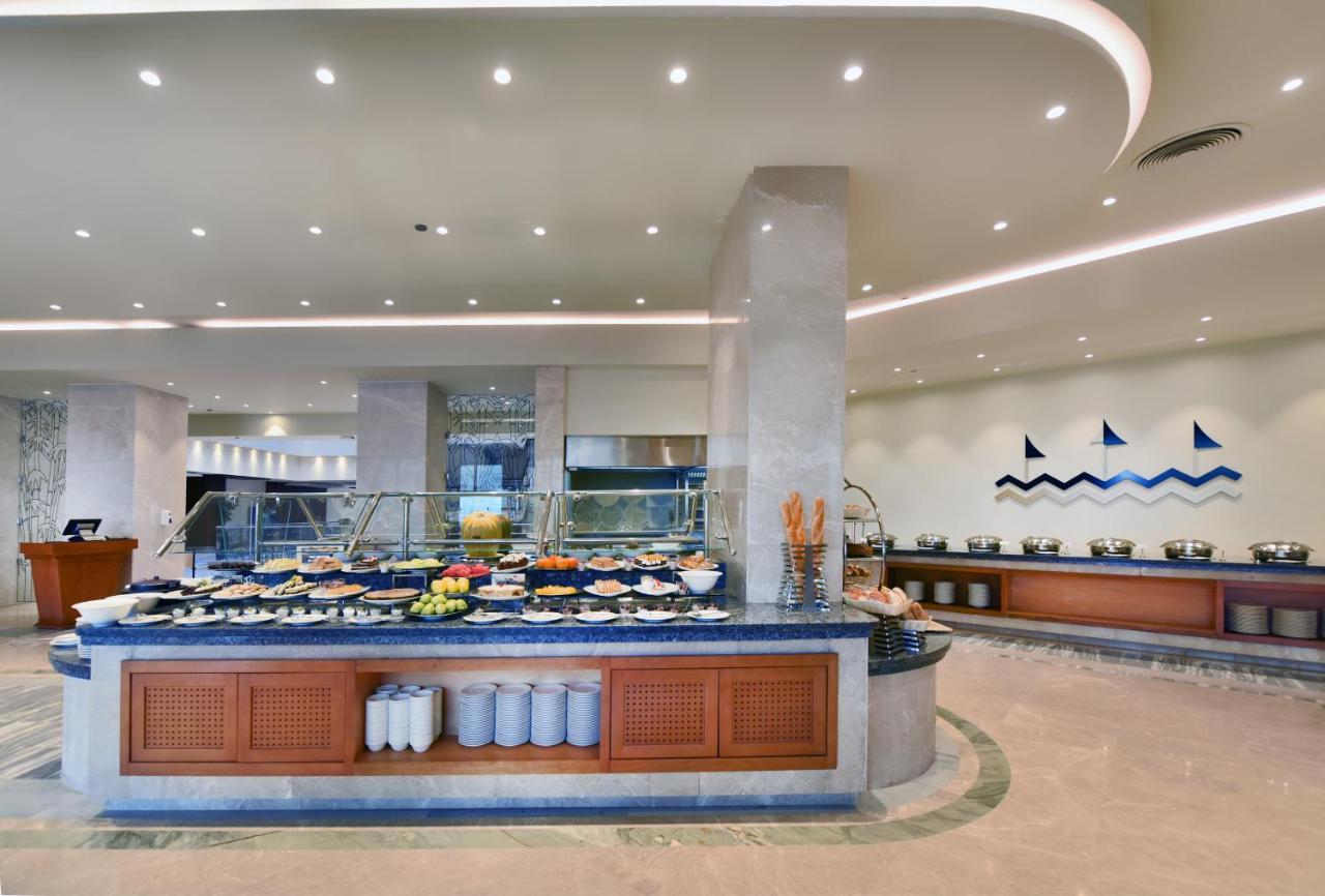 Tolip Resort El Galala Majestic Ain Sukhna Exterior photo The image shows a buffet-style dining area in a restaurant or hotel. There are several food stations with a variety of dishes displayed, including fruits, salads, and other food items. The layout features a large, central island with a wooden base an
