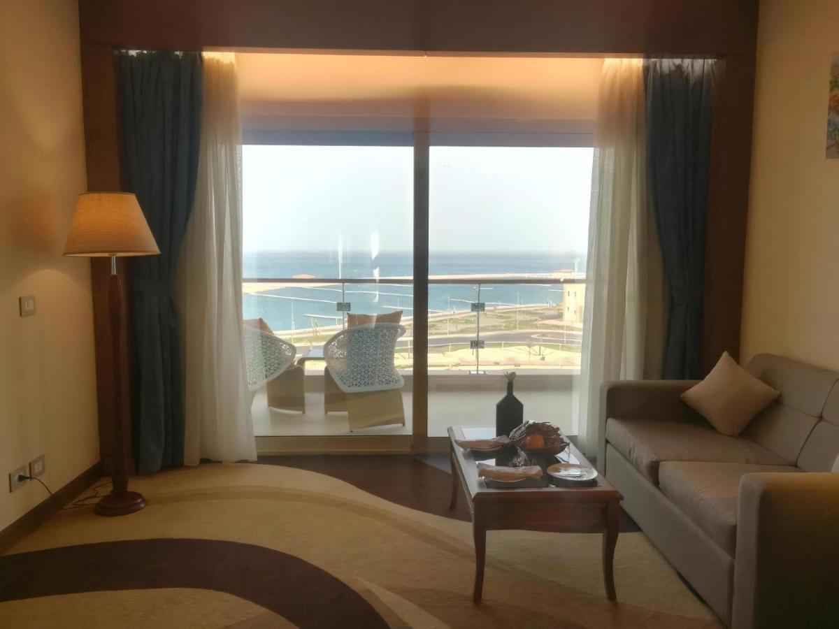 Tolip Resort El Galala Majestic Ain Sukhna Exterior photo The photo shows a stylish interior of a room with a large window overlooking the sea. The window features sheer and thick curtains and is framed by a balcony with two chairs visible outside. Inside, there is a comfortable-looking sofa, a coffee table