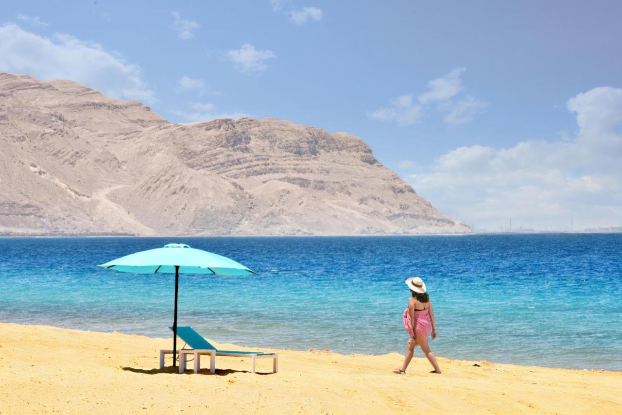 Tolip Resort El Galala Majestic Ain Sukhna Exterior photo The photo depicts a serene beach scene. In the foreground, there is a sandy shore with a single beach chair and an umbrella in a light blue color. A person dressed in a pink outfit is walking along the shoreline, enjoying the beach atmosphere. In the